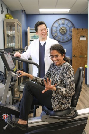 Dr. Lu supervising smiling patient on recumbent exercise bike in rehab gym